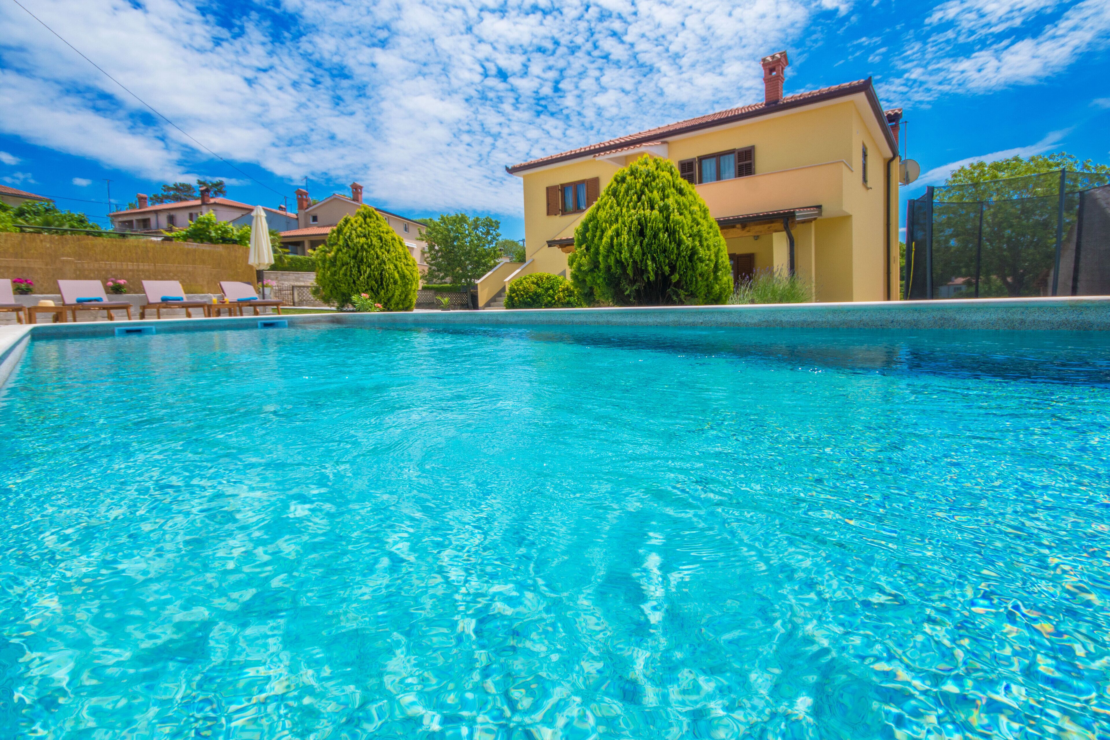 Una piscina con agua azul cristalina, rodeada de sillas de descanso y un edificio amarillo con techo rojo.
