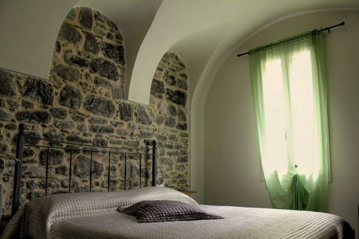 A bedroom with a stone wall, a metal bed frame, a green curtain, and a bed with a quilted blanket and a pillow.