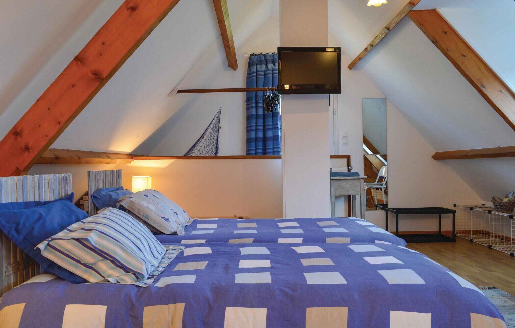 Un dormitorio con techo inclinado, cama con ropa de cama azul y blanca, televisor montado en la pared y un pequeño escritorio.