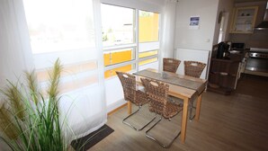 Ein Essbereich mit Tisch und Stühlen, ein Blick durch große Fenster und eine Küche im Hintergrund.