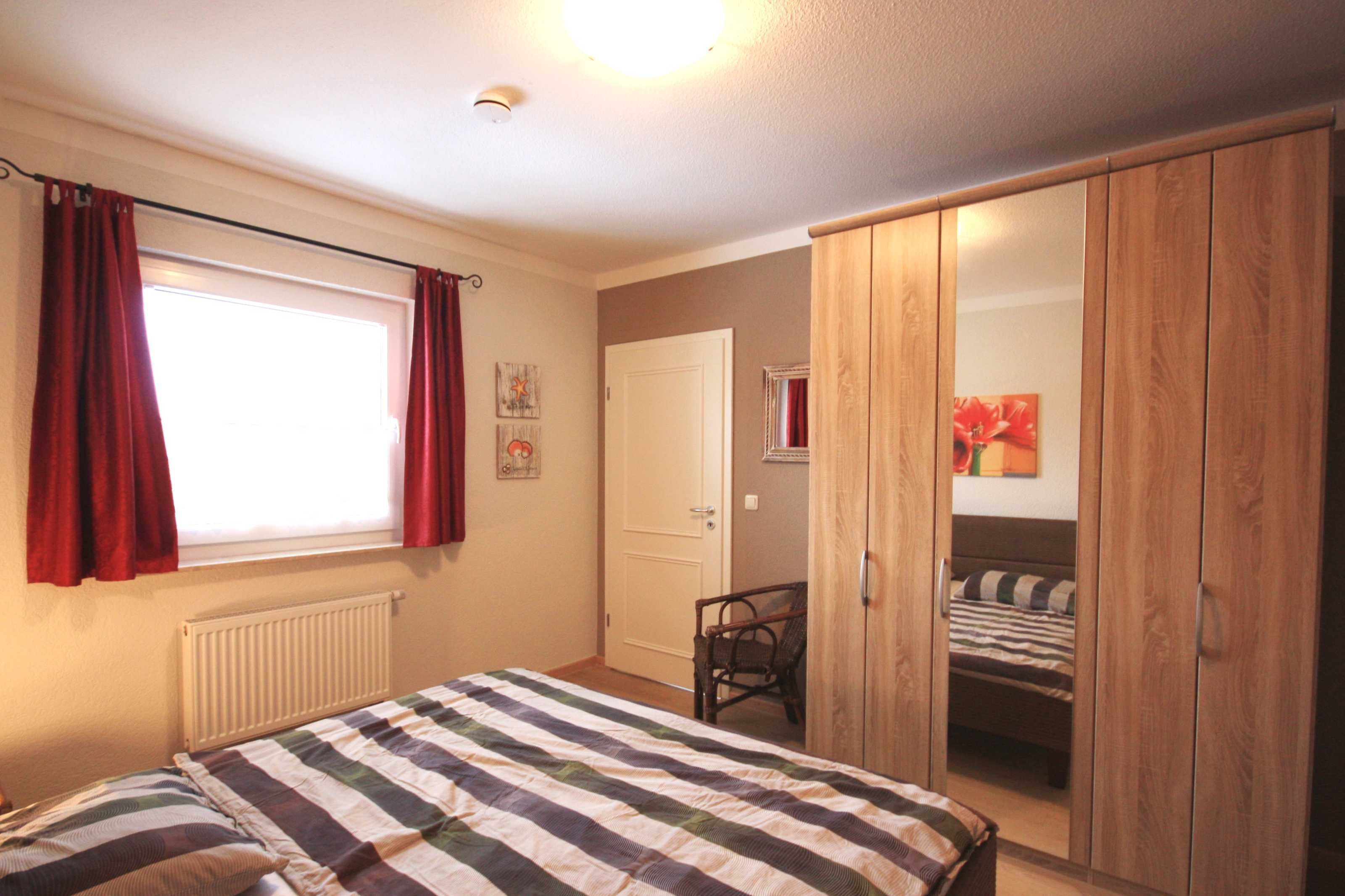 Ein Schlafzimmer mit Bett, Fenster mit Vorhängen, Heizkörper, Tür und Kleiderschrank.