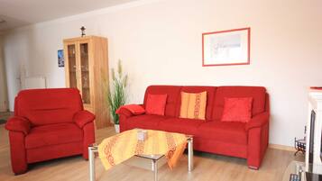 Una sala de estar con un sofá rojo, un sillón rojo, una mesa de centro de vidrio, un aparador de madera y un cuadro enmarcado en la pared.