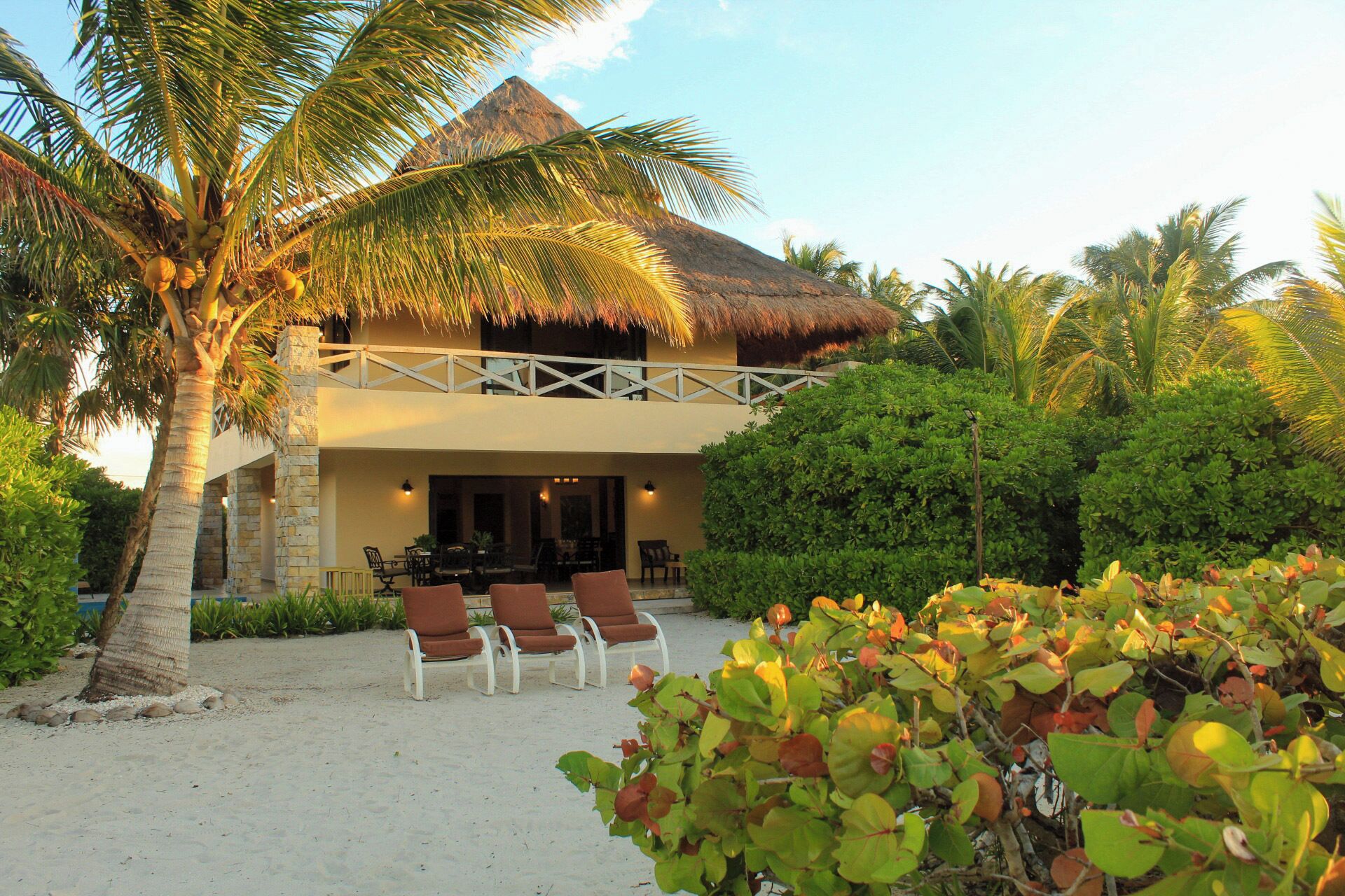 A two-story building with a thatched roof, surrounded by palm trees and a sandy area with lounge chairs.