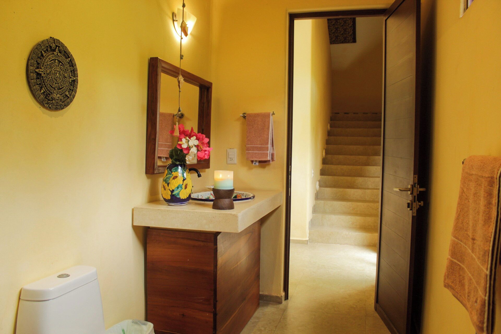 Une salle de bain avec un meuble-lavabo en bois, un miroir, une assiette décorative, un vase avec des fleurs et une bougie allumée.