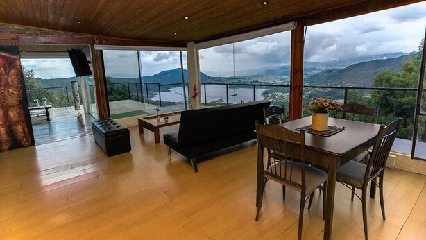 Une pièce spacieuse avec un plancher en bois, un canapé noir, une table à manger avec des chaises et une grande fenêtre donnant sur un lac et des montagnes.
