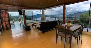 A spacious room with a wooden floor, a black sofa, a dining table with chairs, and a large window offering a view of a lake and mountains. - Hostal San Rafael La Calera (La Calera)