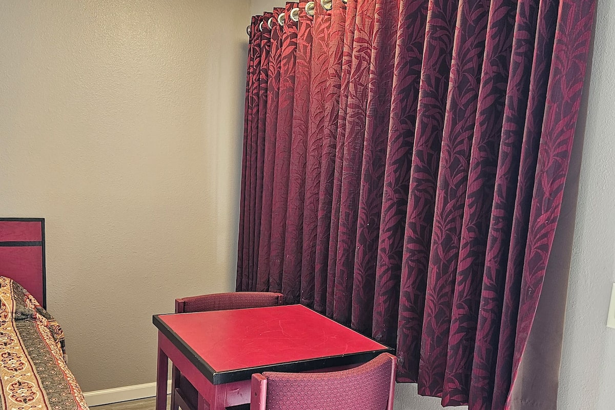 A small room with a single red table and chair, a patterned bed, and a window with dark red curtains.