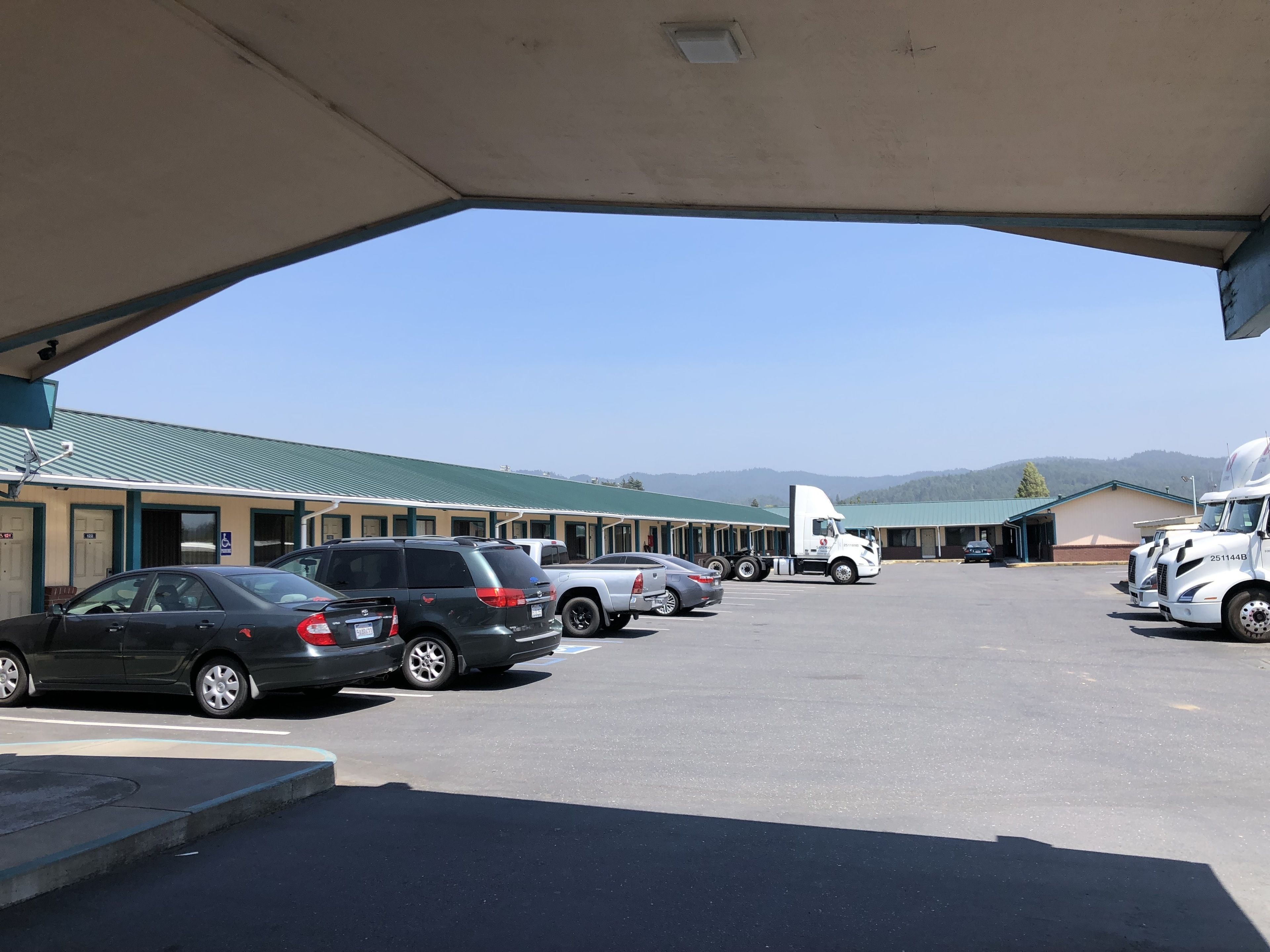 A parking lot with several cars and a large truck, a building with a green roof, and a mountainous backdrop.