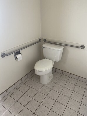 A standard white toilet with a safety bar on the left and right sides, and a roll of toilet paper mounted on the wall.