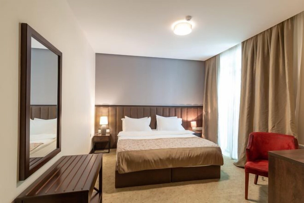 Een hotelkamer met een groot bed, een houten bankje, een rode stoel en een grote spiegel aan de muur.