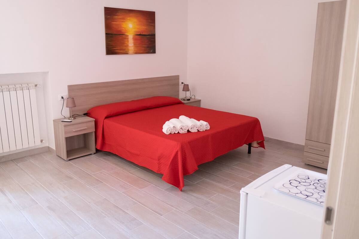 A neatly made bed with a red bedspread and white towels, a nightstand with a lamp, a wall-mounted painting, and a radiator.