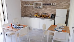 Une cuisine moderne avec des armoires blanches, un dosseret carrelé et un réfrigérateur encastré. On y trouve deux tables à manger avec des chaises blanches, et la cuisine est dotée d’une fenêtre qui laisse entrer la lumière naturelle.
