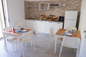 A modern kitchen with white cabinets, a tiled backsplash, and a built-in refrigerator. There are two dining tables with white chairs, and the kitchen has a window providing natural light.