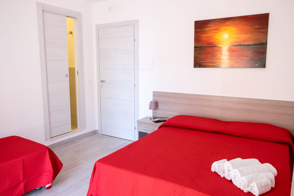 A hotel room with a bed, a bedside table with a lamp, a painting of a sunset, and a door leading to a bathroom.