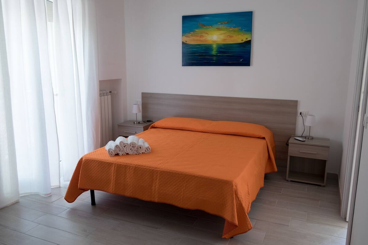 A neatly made bed with an orange bedspread, a bedside table with a lamp, and a painting of a sunset on the wall.