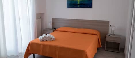 Un lit bien fait, recouvert d’une couvre-lit orange, une table de chevet avec une lampe et un tableau représentant un coucher de soleil accroché au mur.