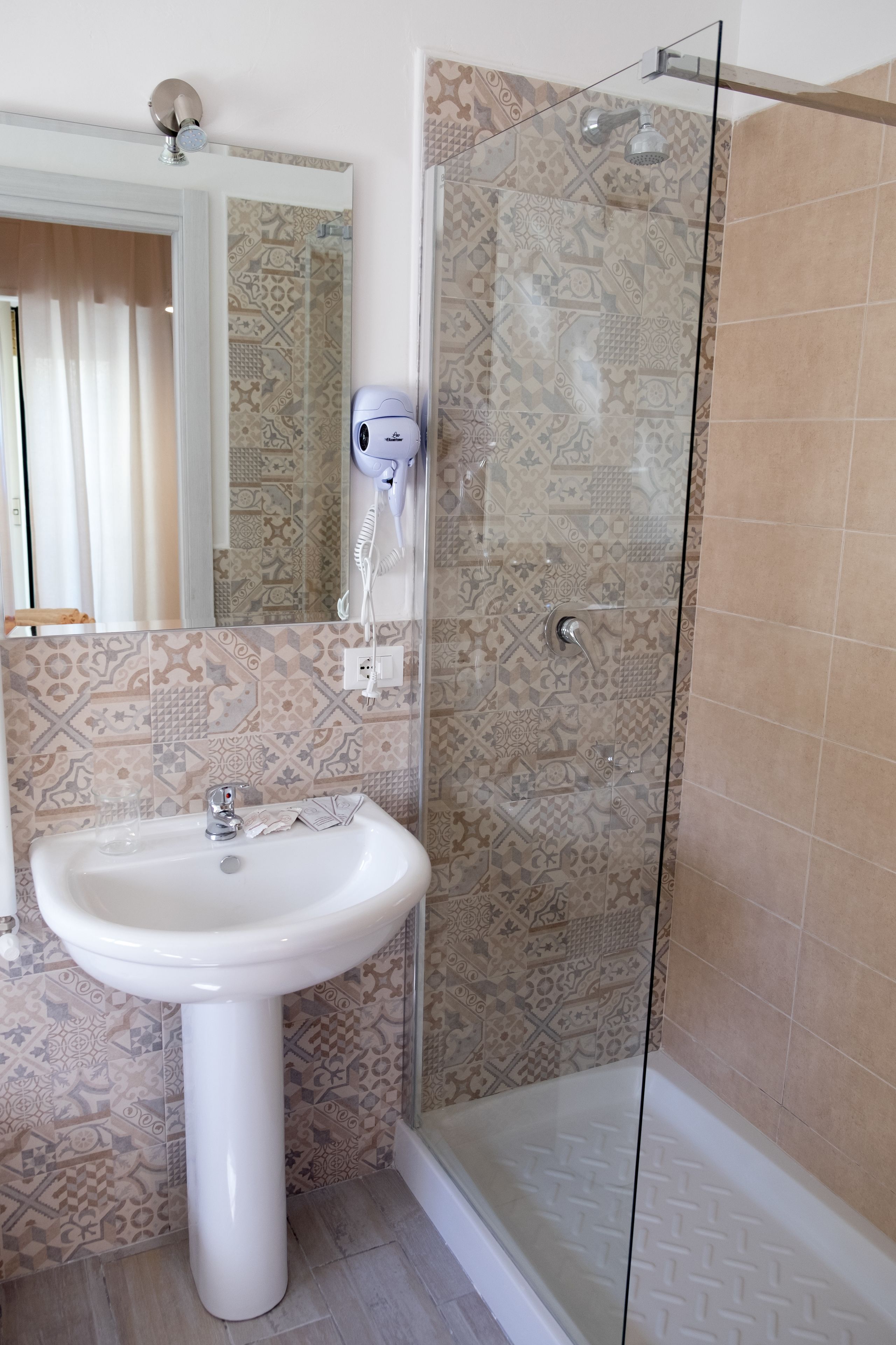 Ein Badezimmer mit weißem Waschbecken, einem Spiegel, einem Fön und einer Dusche mit gemusterten Fliesen.