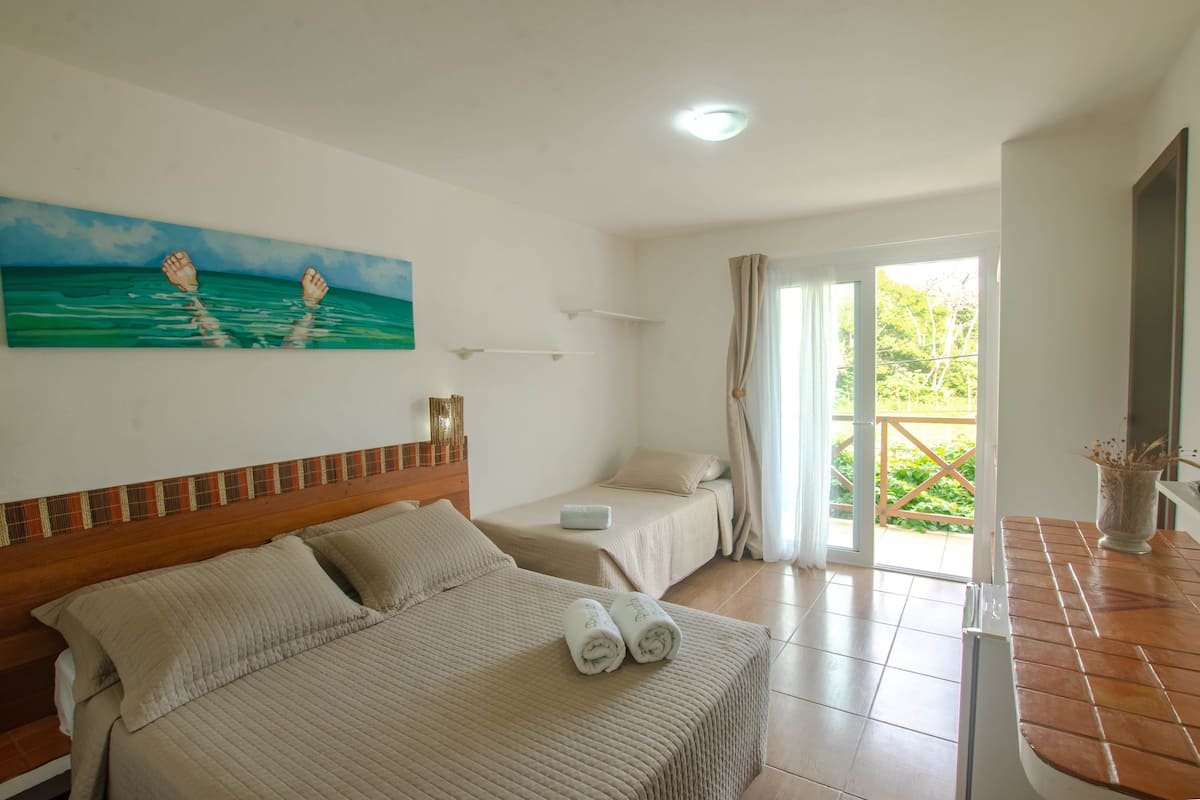 Une chambre d’hôtel avec un grand lit, un plus petit lit, un tableau représentant des pieds dans l’eau, une fenêtre avec des rideaux et une vue sur la verdure à l’extérieur.