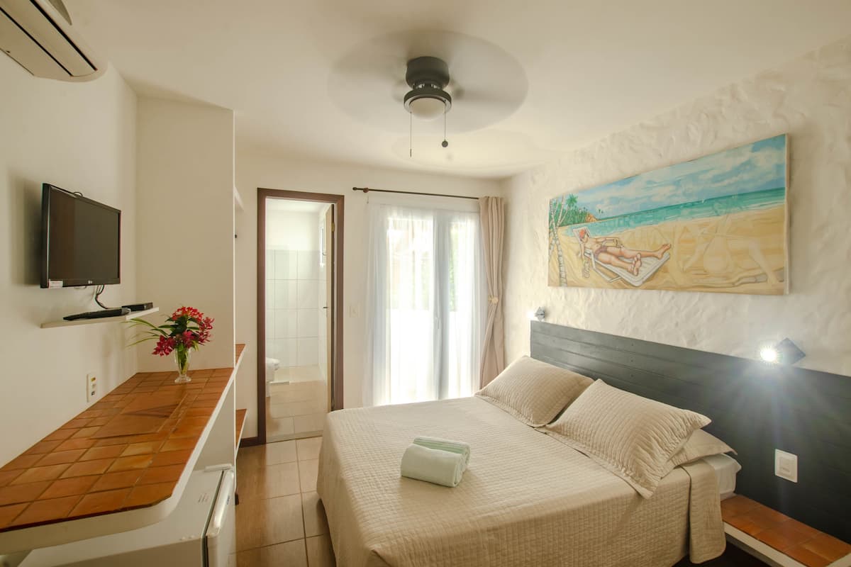 Une chambre d’hôtel avec un lit, une télévision, un tableau représentant une scène de plage et une porte donnant sur une salle de bain.