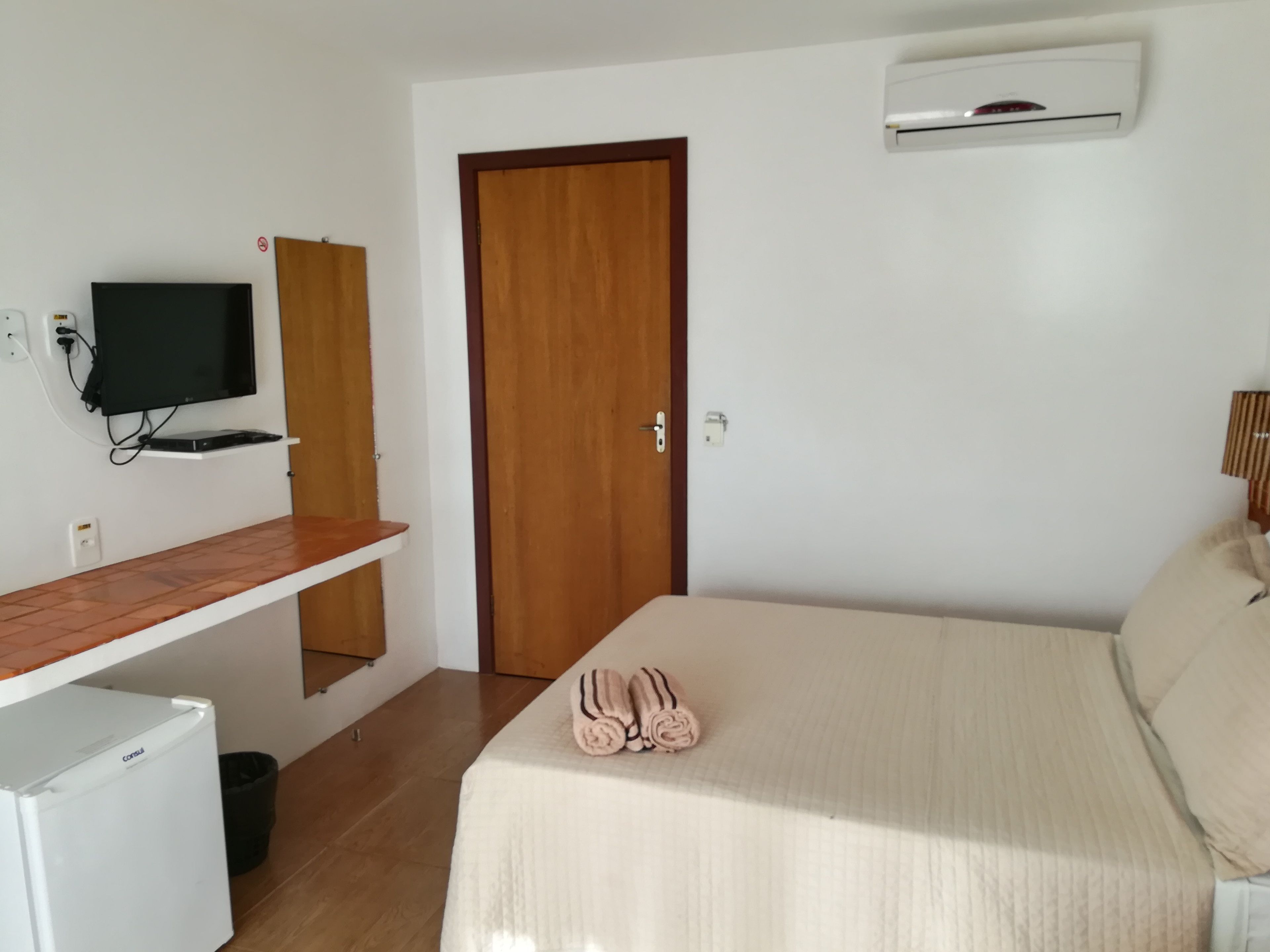 A bedroom with a bed, a TV, a small fridge, a door, and an air conditioner.