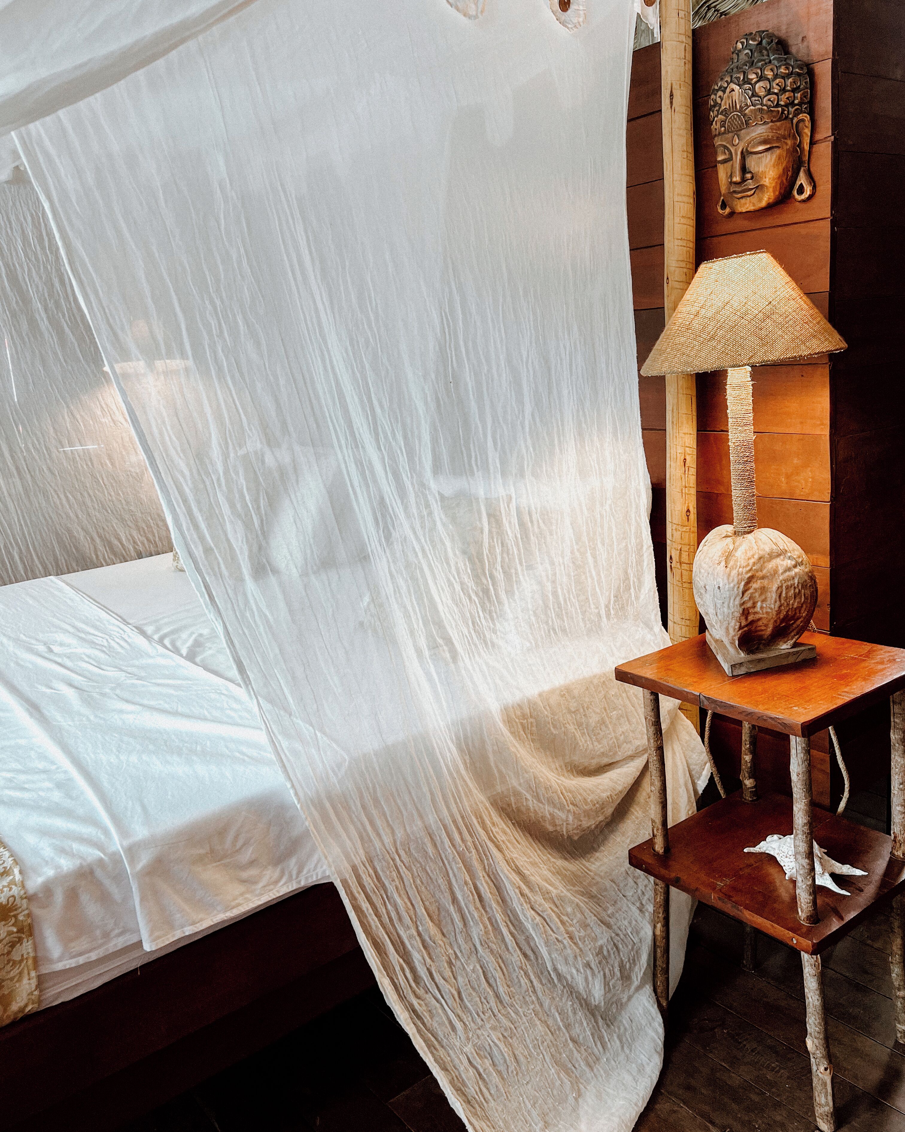 A bed with a mosquito net, a wooden nightstand with a lamp, and a Buddha statue on the wall.
