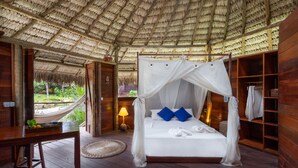 Ein Zimmer mit einem Bett unter einem Reetdach, einem Holztisch und Blick ins Freie.