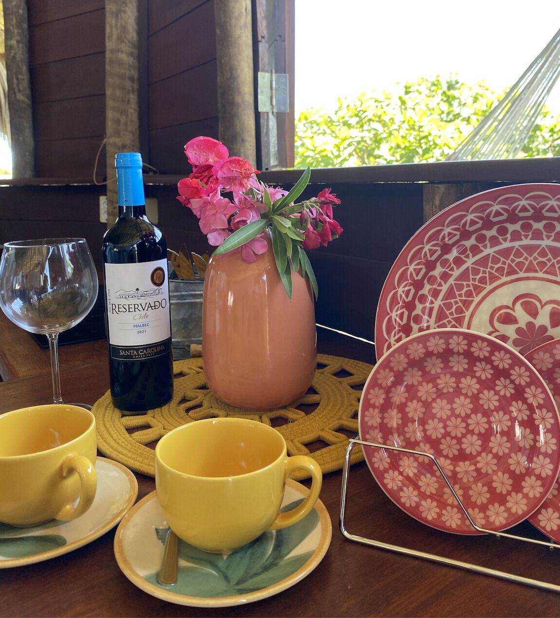 A table with a bottle of wine, two yellow cups, a vase with pink flowers, and a decorative plate rack.