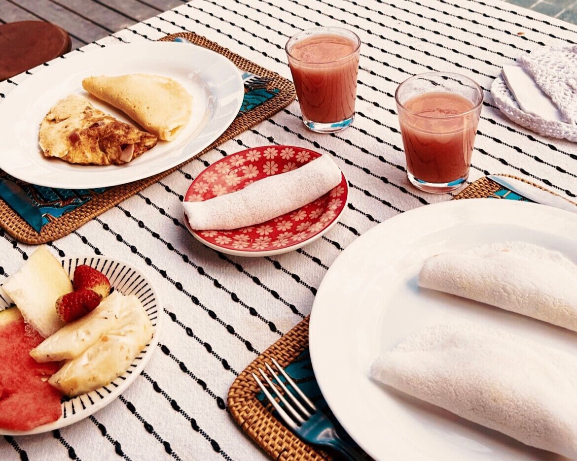 Una mesa con crepas, un tazón de fruta y dos vasos de una bebida rosada.