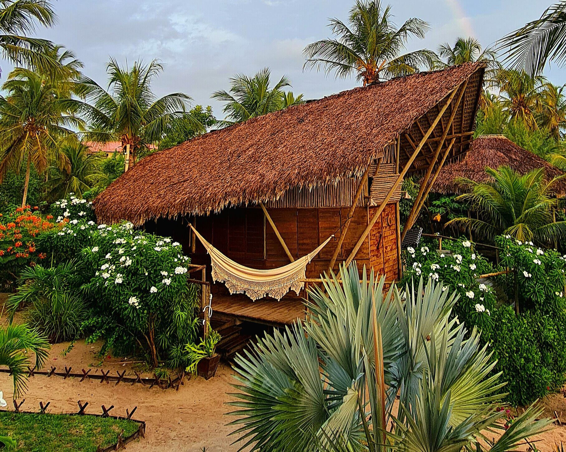 A thatched-roof hut with a hammock, surrounded by tropical plants and palm trees.
