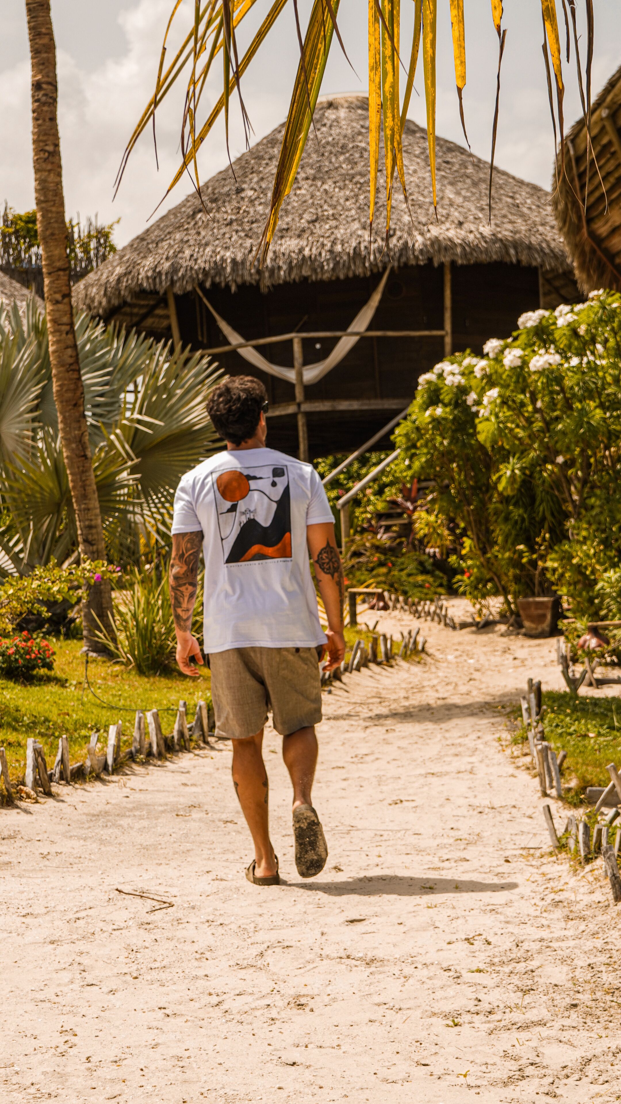 Un homme marchant sur un sentier sablonneux en direction d’une construction au toit de chaume, entourée d’une végétation tropicale.