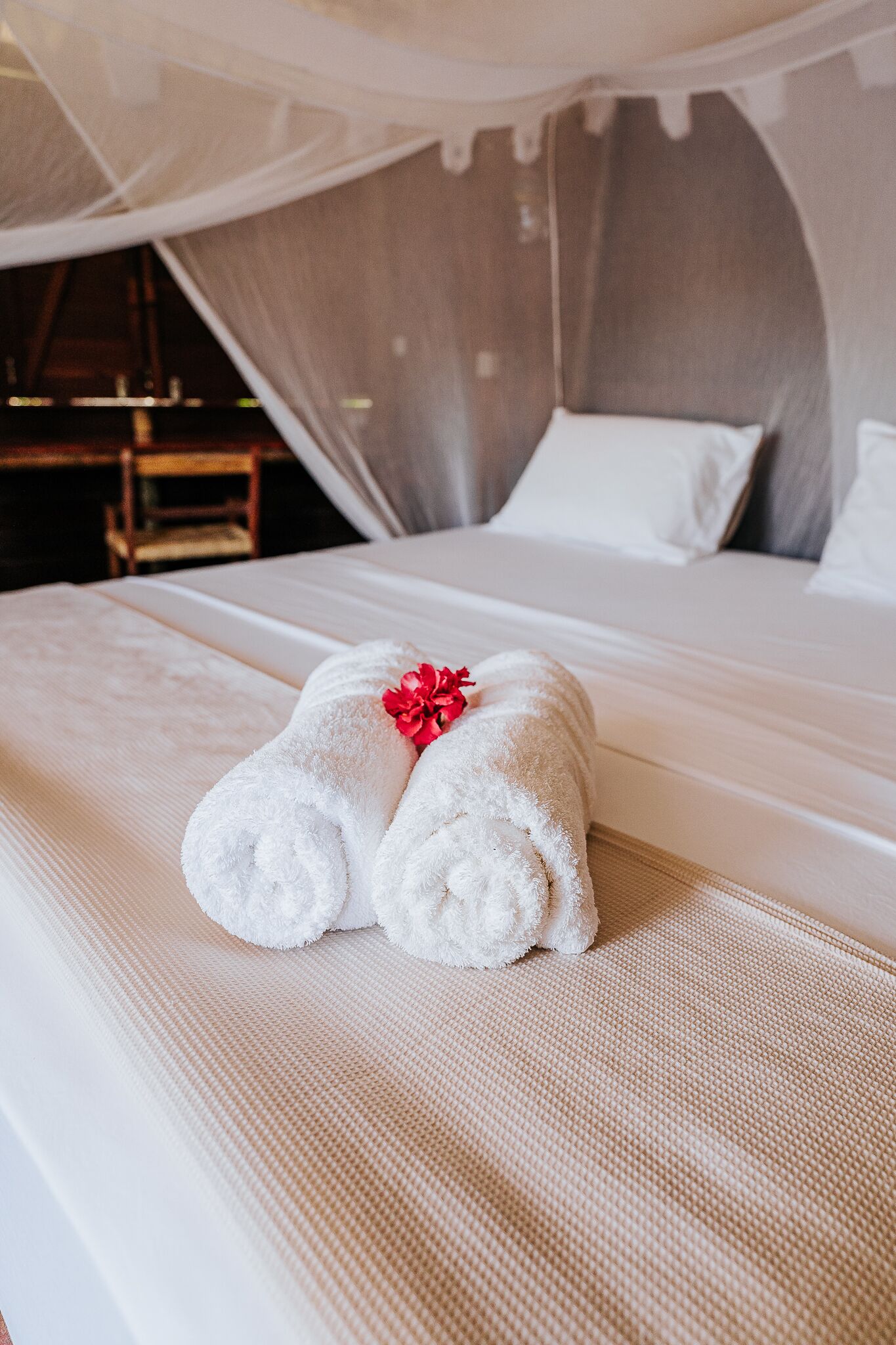Un lit bien fait, avec du linge de lit blanc et une fleur rouge, dans une pièce où se trouve un lit à baldaquin et, au fond, une chaise en bois.