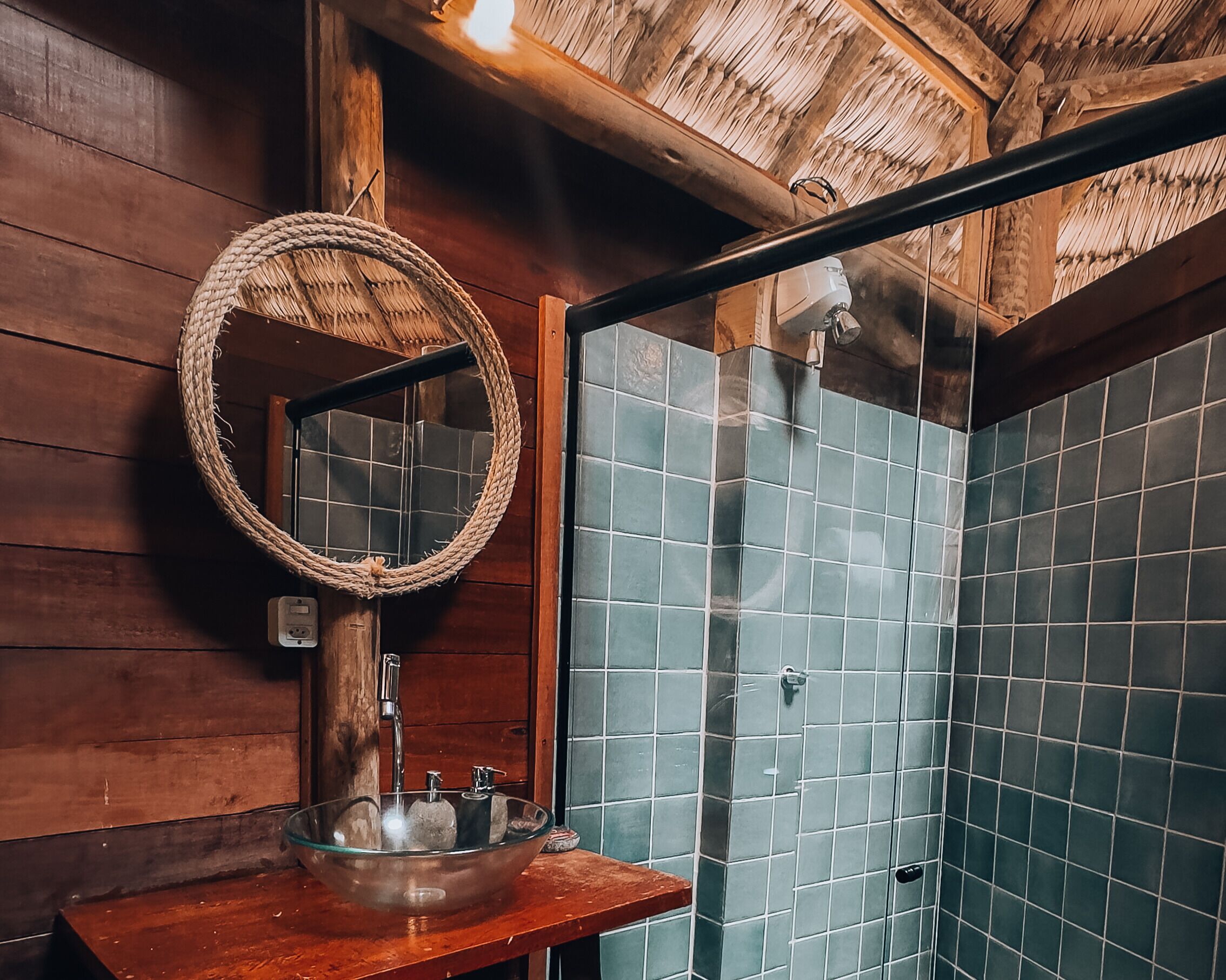 Une salle de bain avec un plafond en bois, un lavabo en forme de bol en verre, un miroir à motifs tissés et une douche aux carreaux bleus.