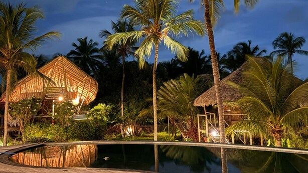 Ein Poolbereich eines Resorts bei Nacht mit Holzdeck, strohgedeckten Gebäuden und Palmen.
