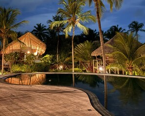 Área de piscina de un resort durante la noche, con muelle de madera, estructuras de techo de paja y palmeras.