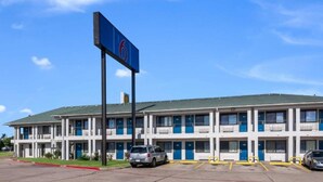 Un motel avec une enseigne bleue indiquant le numéro de chambre 6. Il s’agit d’un bâtiment à deux étages comprenant plusieurs chambres, chacune dotée d’un balcon. Deux voitures sont garées devant.