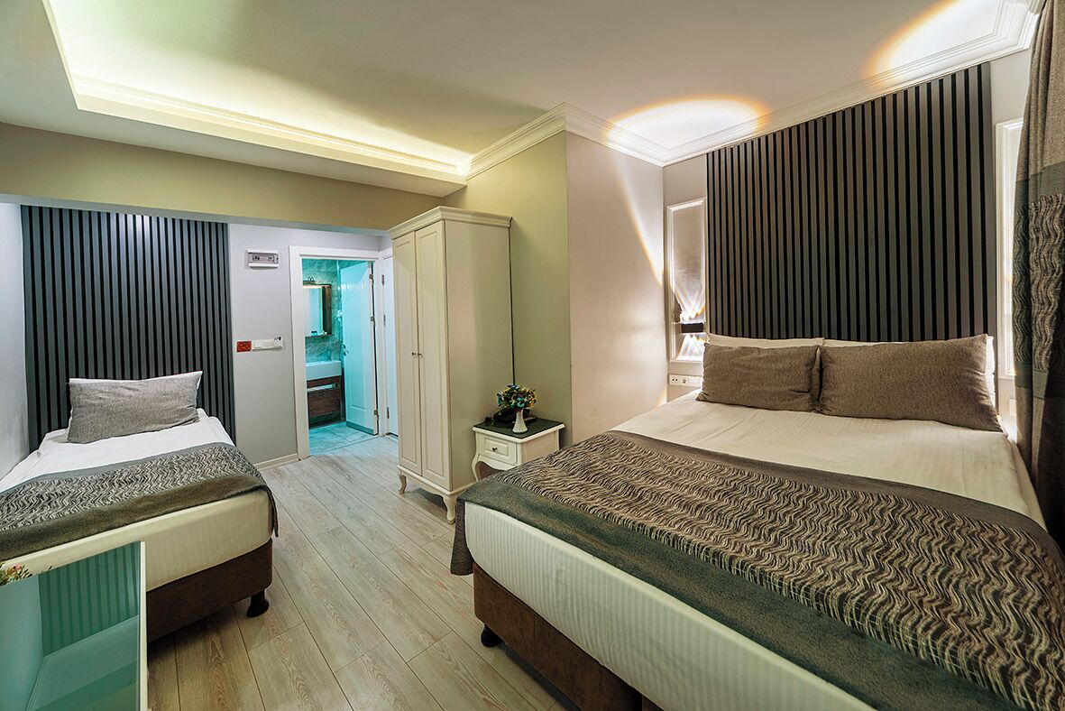 Habitación de hotel con dos camas, un armario, una mesita de noche con lámpara y un baño visible a través de una puerta abierta.