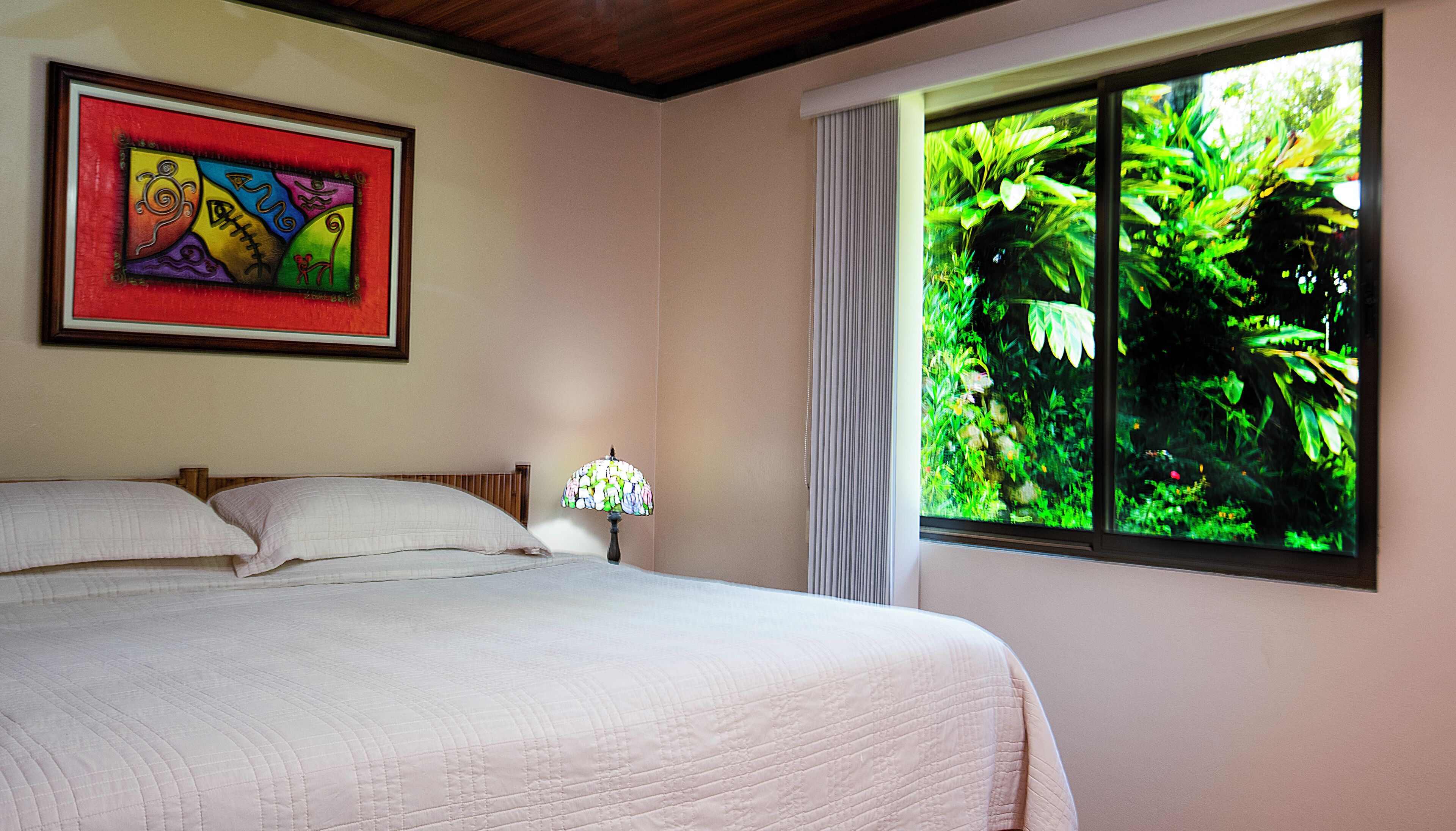 A bedroom with a bed, a nightstand, a lamp, a window with a view of greenery, and a colorful painting on the wall.