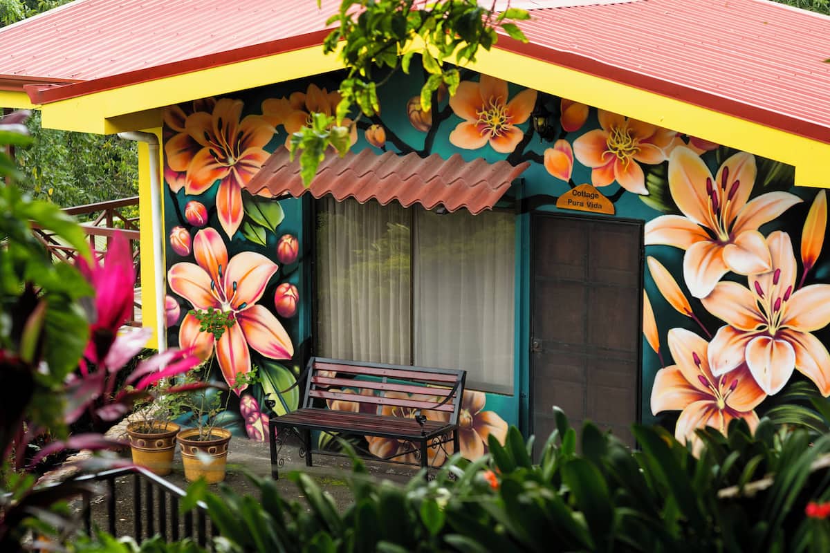 Een kleurrijk gebouw met een levendige bloemenmuur, een bankje en een deur met de tekst 'Cottage Pura Vida'.