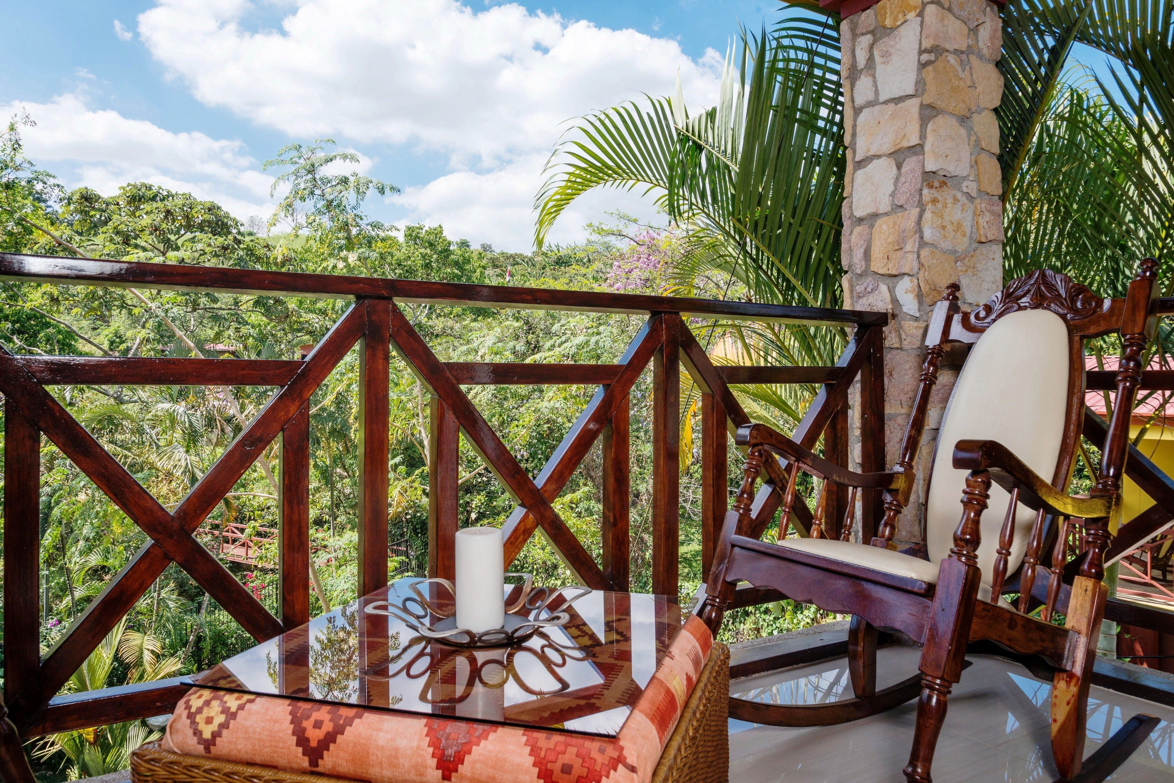 A wooden deck with a stone pillar, a wicker chair, and a cushioned bench with a decorative pillow and candle.
