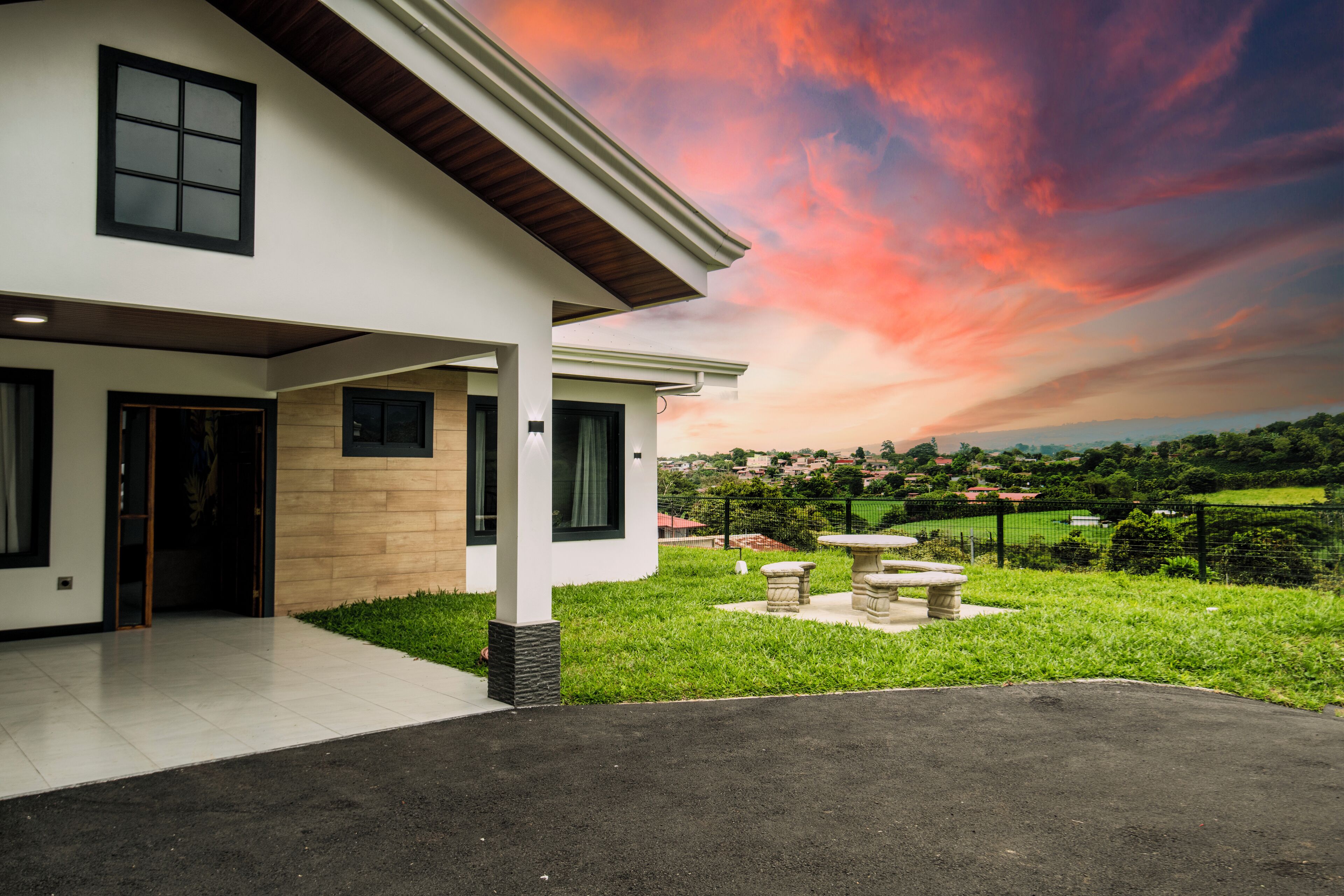 A modern two-story house with a covered porch, a stone bench, and a view of a green landscape.