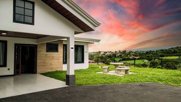 A modern two-story house with a covered porch, a stone bench, and a view of a green landscape.