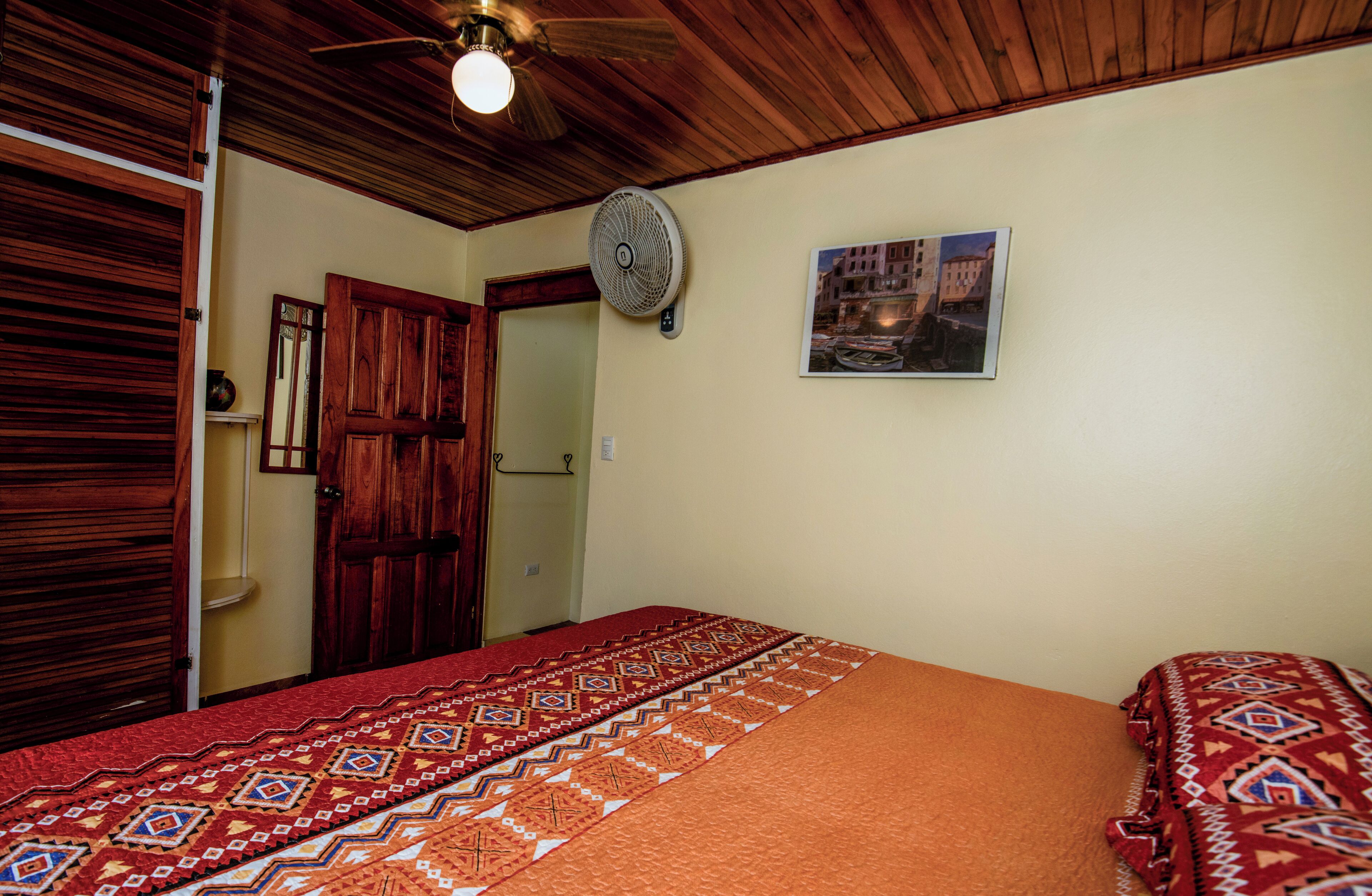 A bedroom with a wooden ceiling, a bed with a patterned bedspread, a wall-mounted fan, and a framed picture on the wall.
