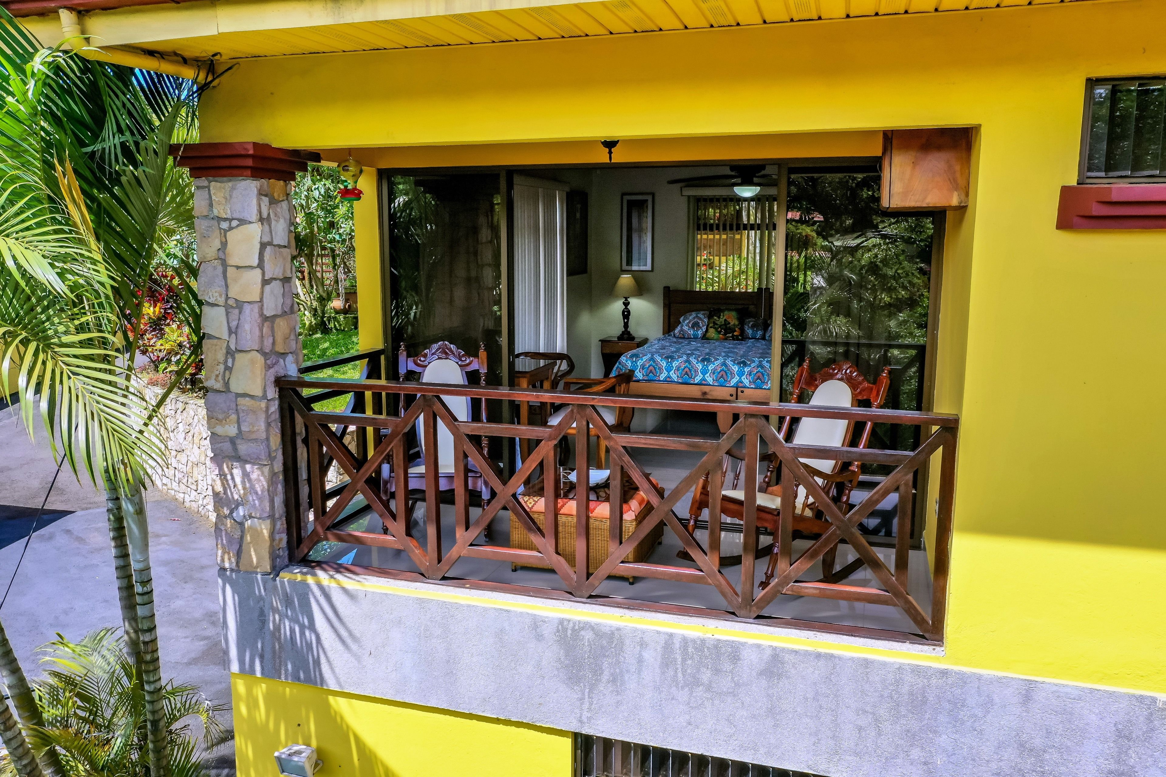A balcony with a stone pillar, a bed with a blue patterned bedspread, and a nightstand with a lamp.