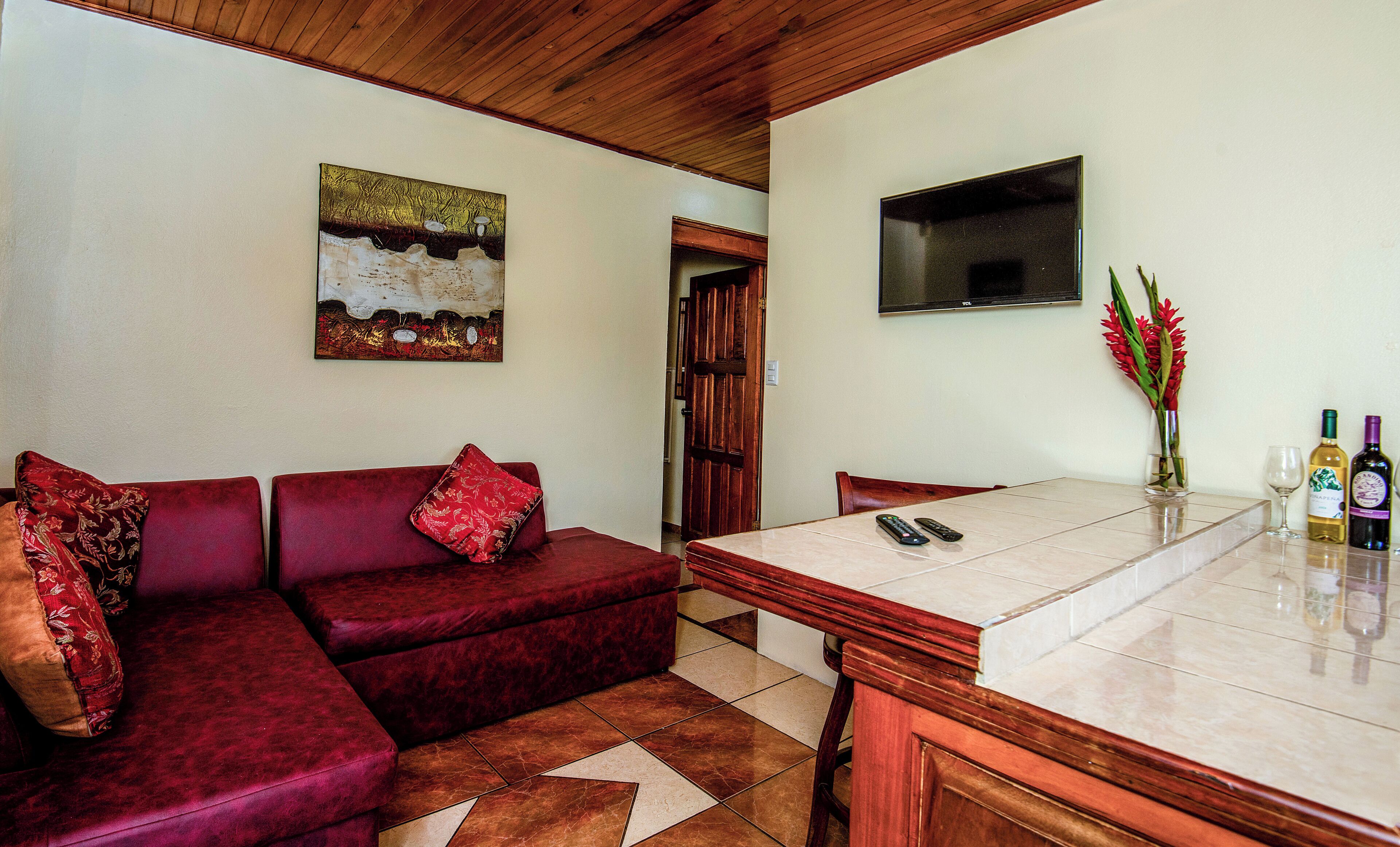 A living room with a red sofa, a painting on the wall, a TV mounted above a wooden table, and a door leading to another room.