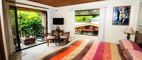 Une chambre dotée d’un grand lit, d’une télévision, d’un balcon offrant une vue sur la verdure, ainsi que d’un coin repas avec des chaises et une table.