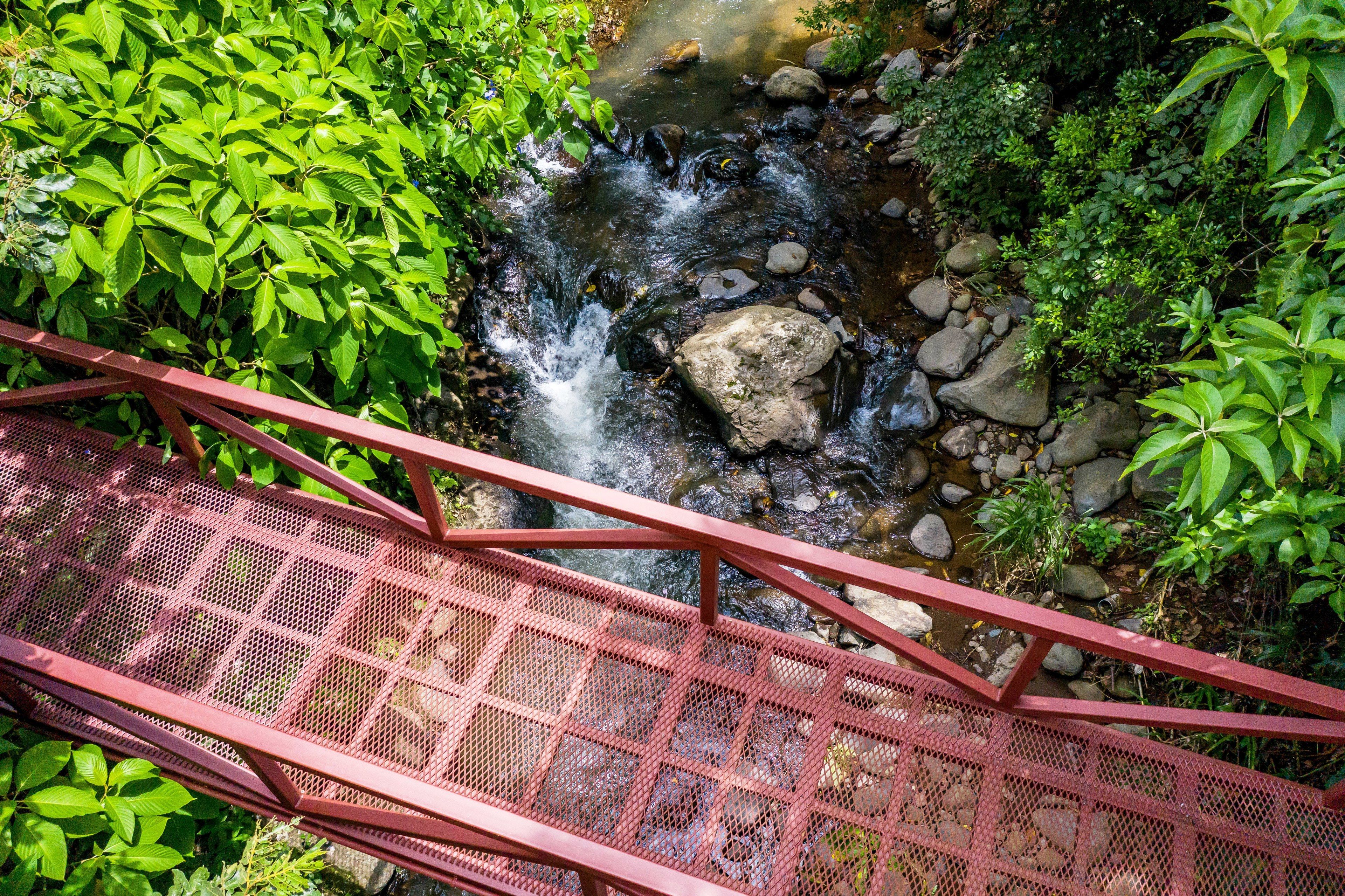 A red metal bridge over a stream with rocks and greenery on either side.