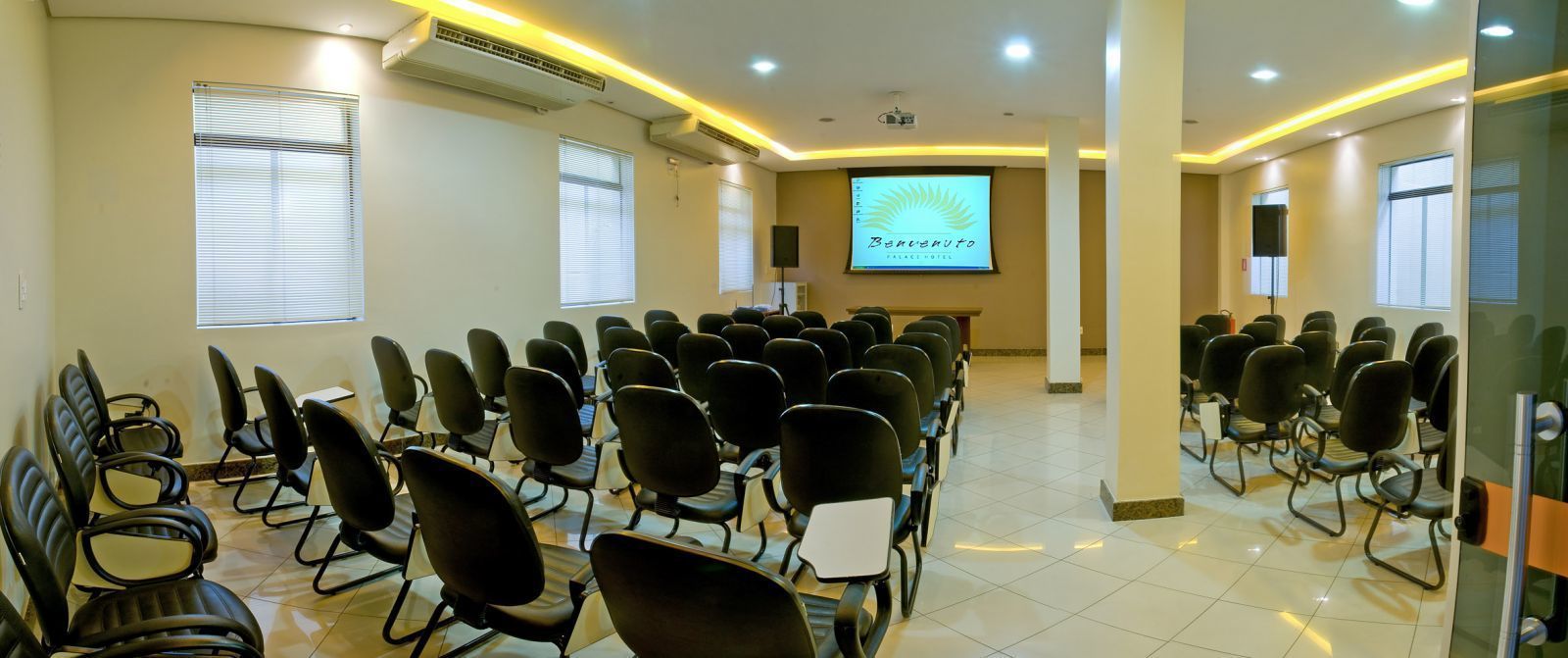 Un salón de conferencias con filas de sillas, una pantalla de proyector y una presentación en la pared.