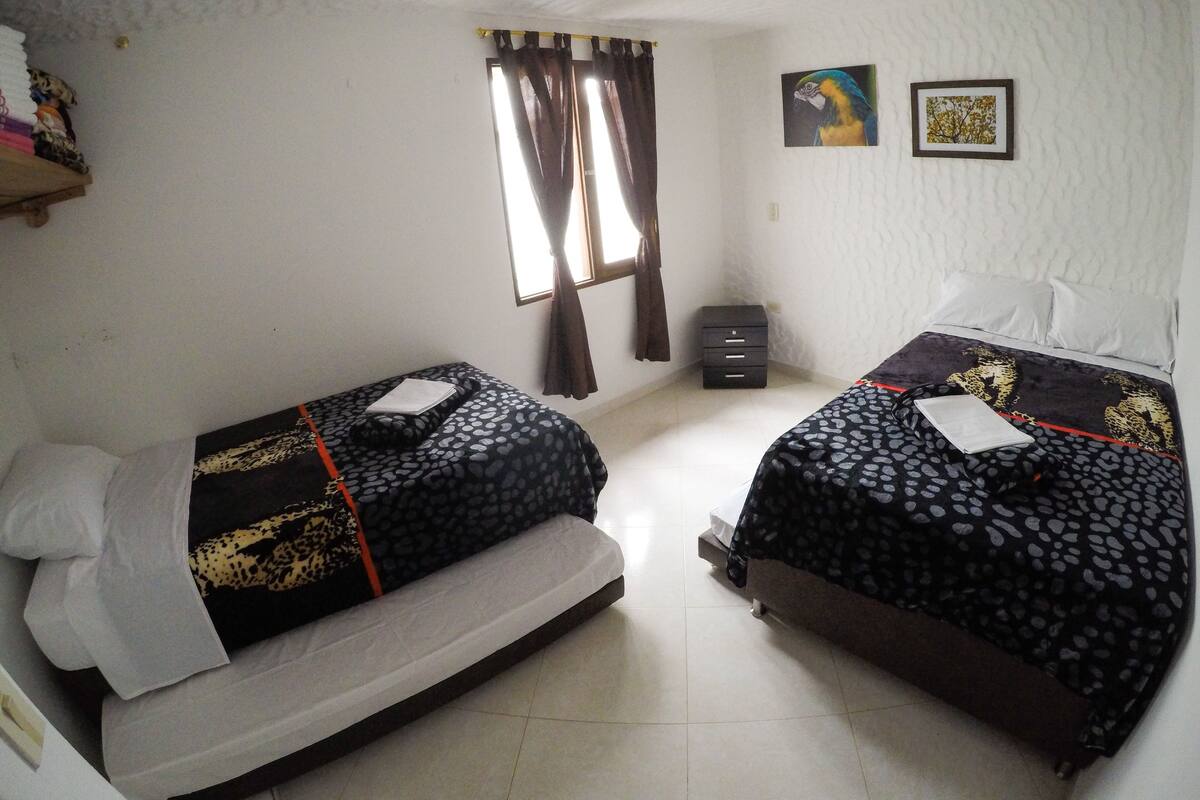 A bedroom with two beds, a window with curtains, a small cabinet, and two framed pictures on the wall.