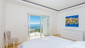 A bedroom with a bed, a chair, a white dresser, a painting, and a balcony with a view of the sea. - Villa Sv. Petar (Opatija)