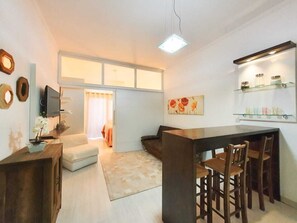 A modern living room with a sofa, a coffee table, and a TV. There is a kitchen area with a bar and chairs.